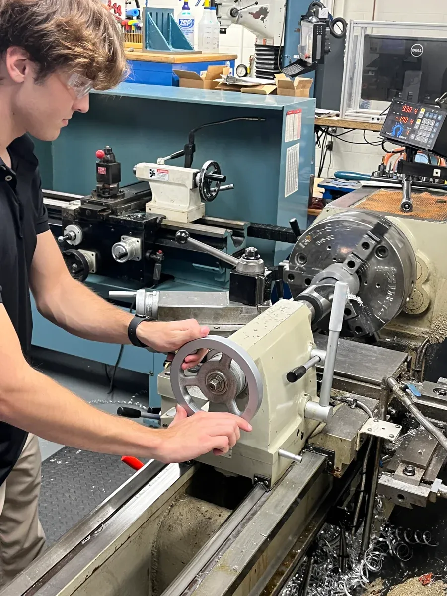 A team member at a manual lathe with one hand on the tailstock handwheel.